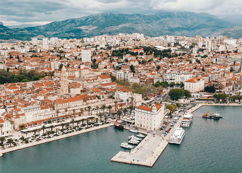 Aerial daytime view of Split, Croatia, highlighting the waterfront Riva promenade, the historic Diocletian's Palace area with its bell tower, and mountains in the background.