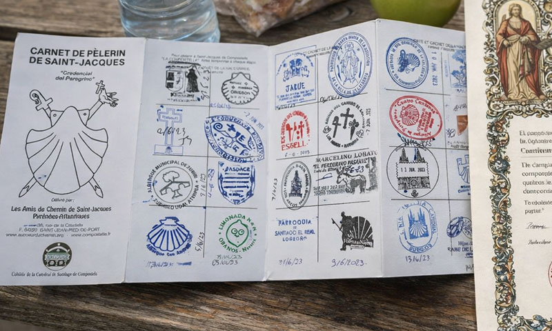 An open Camino de Santiago pilgrim passport filled with ink stamps, sitting on a wooden picnic table next to a lunch sandwich, an apple, and the official Compostela certificate.
