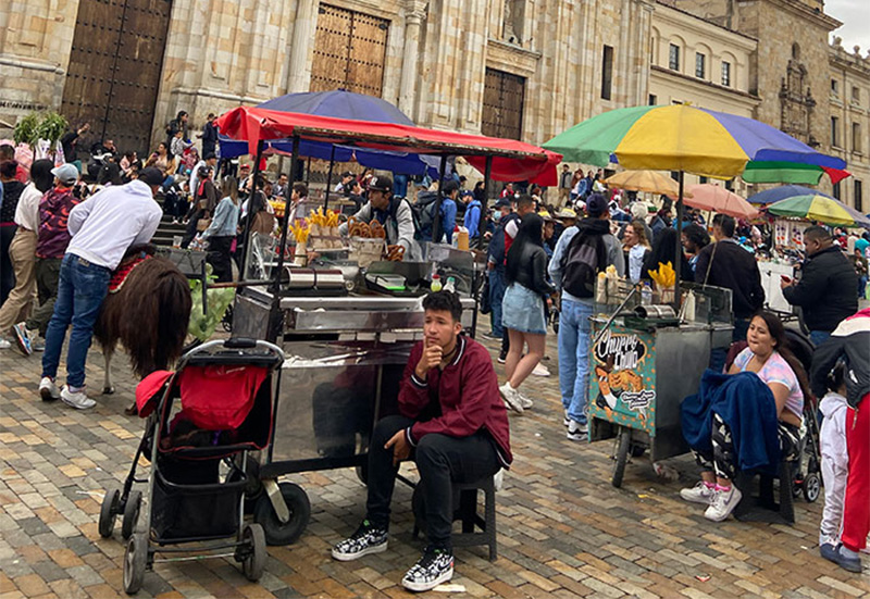 A lively scene in Bogotá’s Plaza de Bolívar featuring several colorful street food carts with red and blue umbrellas, surrounded by locals and tourists against the backdrop of the city’s historic stone buildings.