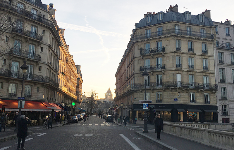 A perspective view down a classic Parisian street at sunset, showing Haussmann-style buildings, cafe awnings, and the warm glow of the evening sun hitting the facades.