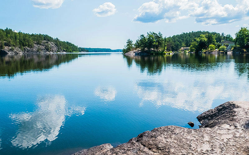 A calm, glassy lake in Sweden reflecting the surrounding green forest and blue sky during a peaceful summer afternoon.