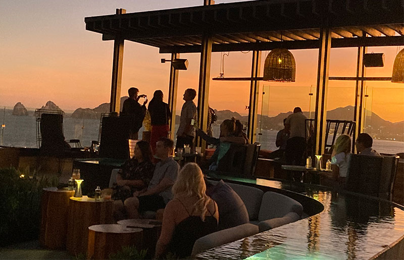 People relaxing with drinks at an open-air rooftop bar under a modern wooden pergola, with a vibrant orange sunset over the ocean in the background.
