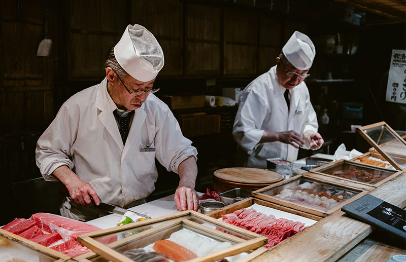 Two sushi chefs in white uniforms and traditional hats prepare fresh nigiri at a wooden counter in a dimly lit Tokyo restaurant.