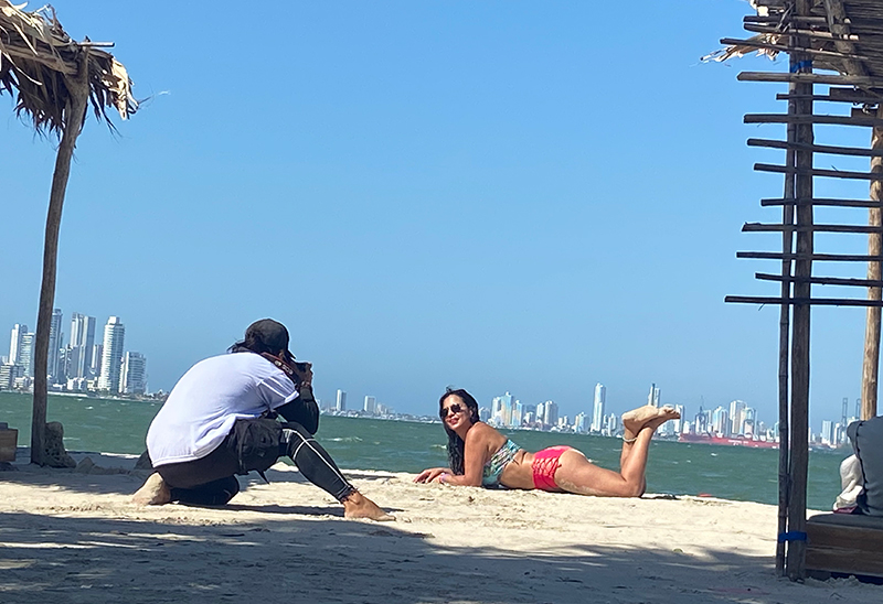 A woman in a bikini posing on a beach while a professional photographer with gear takes her photo.