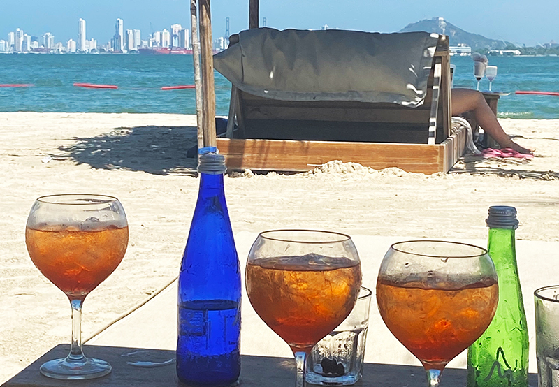 A tropical cocktail in a glass sitting on a wooden table with the beach and ocean blurred in the background.