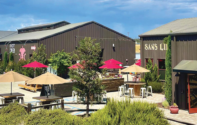 A modern industrial tasting room in Tin City Paso Robles with outdoor seating, umbrellas, and corrugated metal buildings under a bright blue sky.