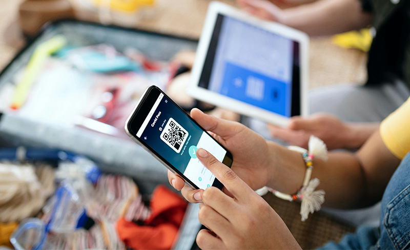 A person holds a smartphone displaying a QR code for a travel document, with a laptop and travel gear like an open suitcase visible in the blurred background of a living room.