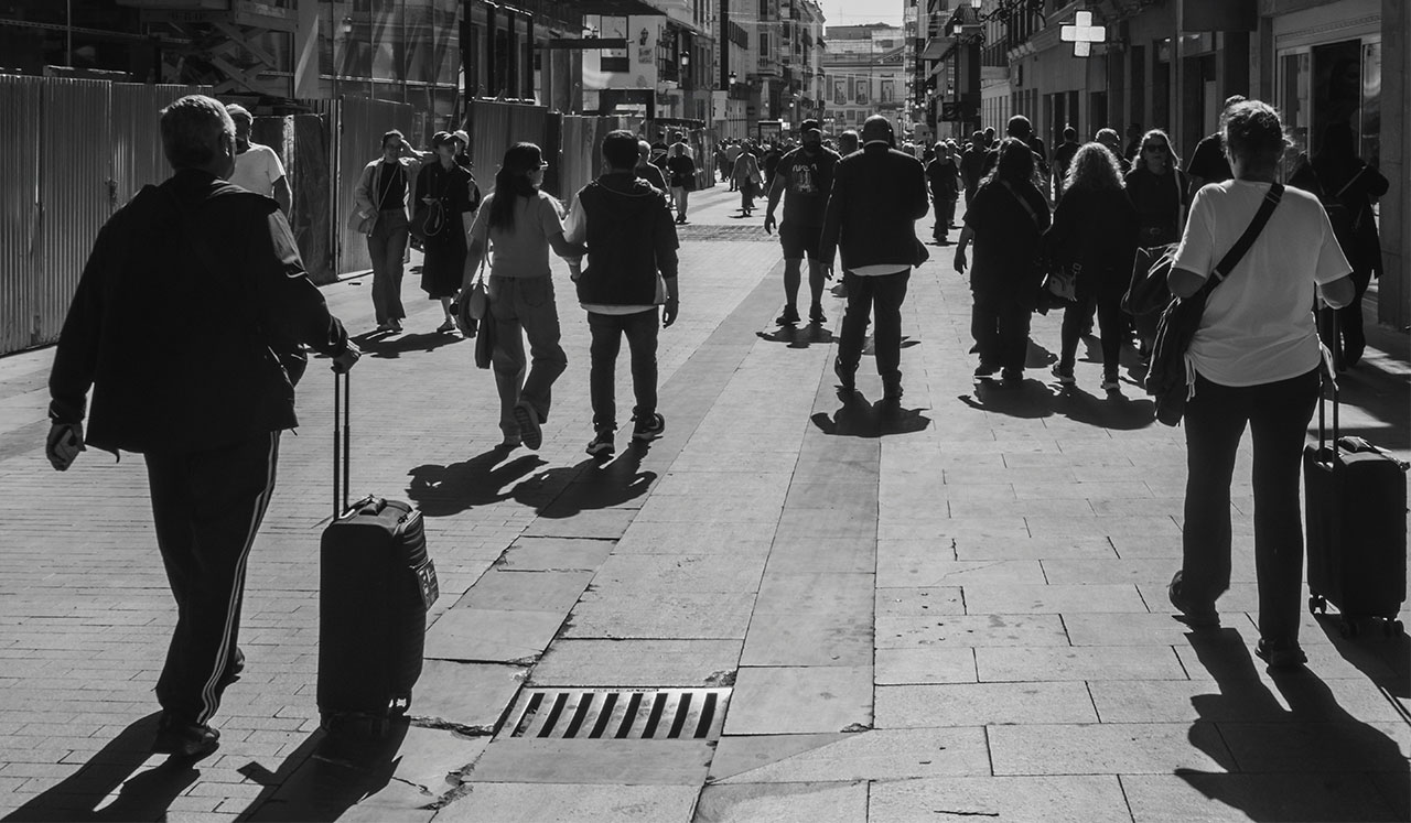 A high-angle, black and white photo of people walking along a wide, sunlit pedestrian street in a European city, with long shadows stretching across the pavement.