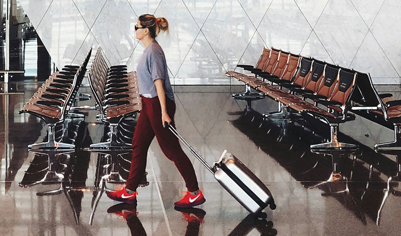 traveler wearing a white top, black leggings, and bright red sneakers pulls a silver carry-on suitcase through a bright, modern airport terminal past rows of empty seating.