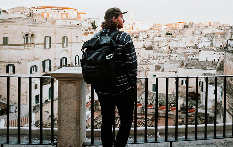 A traveler stands with their back to the camera, looking out over a historic European cityscape under a bright sky. They are wearing a black and white striped shirt and a large black travel backpack. They are leaning against a black metal railing that overlooks terracotta rooftops and a large dome in the distance.
