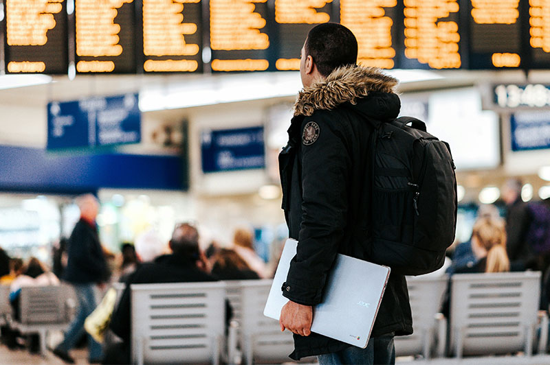 A traveler seen from behind, wearing a black winter parka and carrying a white document.