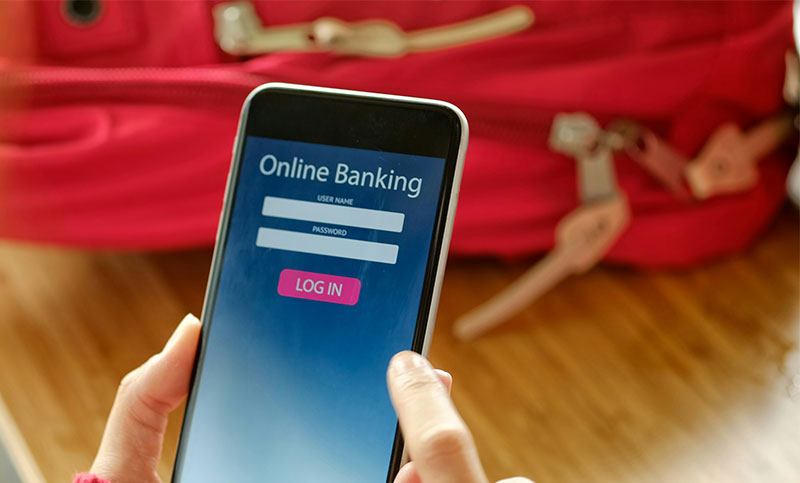 A person holding a smartphone displaying an online banking login screen with a blurred background of travel gear and a passport.