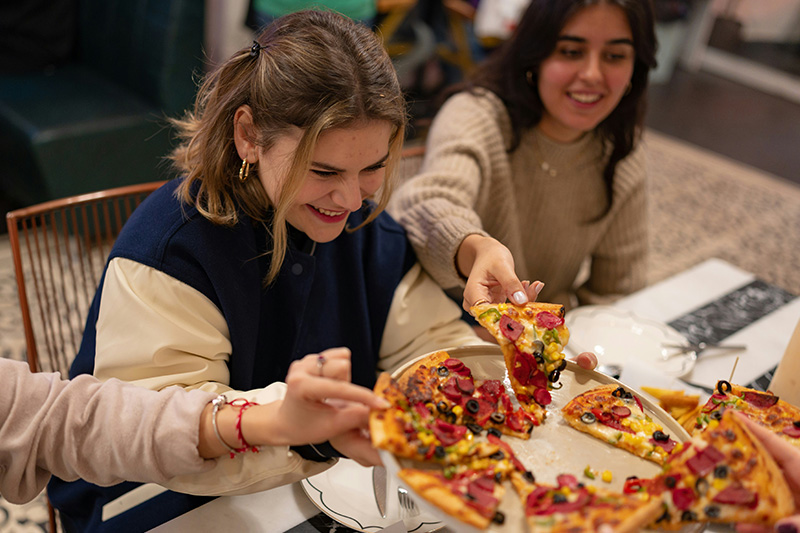 A group of travelers sitting at a wooden table, smiling and laughing as they share a large, fresh pizza in a cozy, rustic restaurant setting.