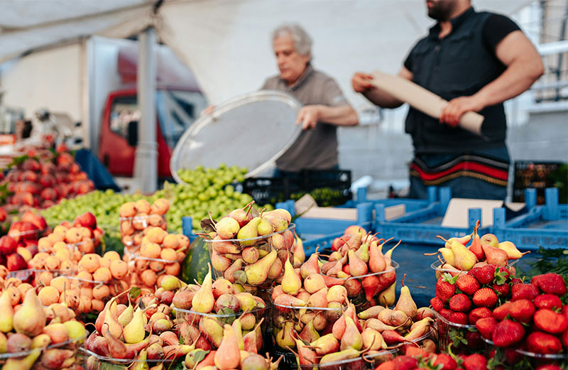 A close-up of a vibrant market stall filled with containers of small pears and strawberries, with two vendors working in the blurred background.