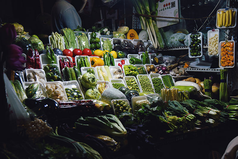 A vibrant night market stall displays rows of fresh vegetables, including peppers, greens, and corn, individually wrapped in trays.