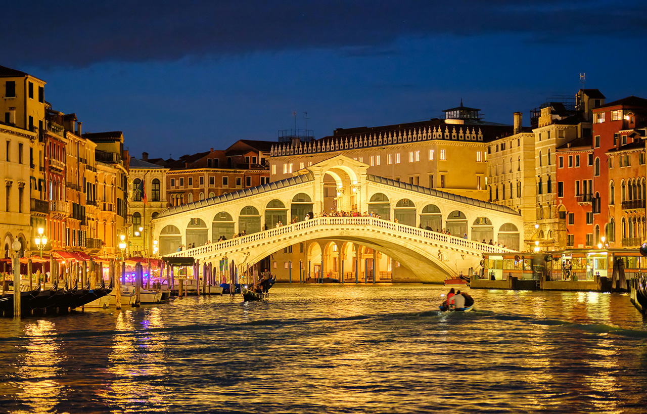 A wide-angle sunset view of the stone Rialto Bridge spanning the Grand Canal in Venice, Italy, with historic buildings lining the water and small boats docked along the bank