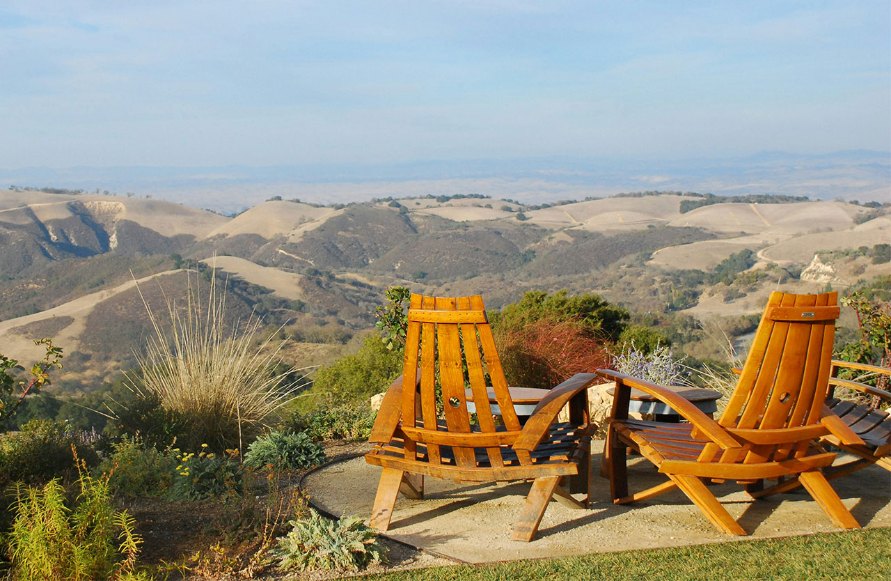 A scenic view from a terrace at DAOU Vineyards in Paso Robles, featuring two empty wooden Adirondack chairs overlooking rolling hills of vineyards under a clear, bright sky.