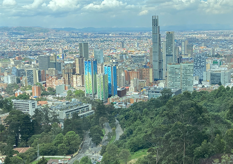 high-altitude, panoramic view from the summit of Cerro de Monserrate looking down over the vast, dense urban sprawl of Bogotá, with the city's skyscrapers and brick buildings stretching toward the horizon under a soft, hazy sky.
