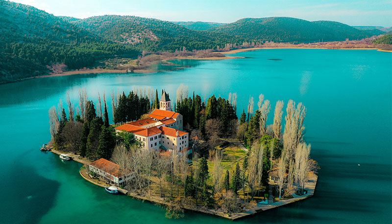 Aerial view of the small Visovac Island featuring a terracotta-roofed monastery surrounded by tall cypress trees in a vibrant blue lake.