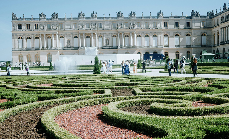 A high-angle view of the manicured Latona Fountain and the expansive geometric gardens at the Palace of Versailles, featuring green lawns and distant pathways under a bright sky.