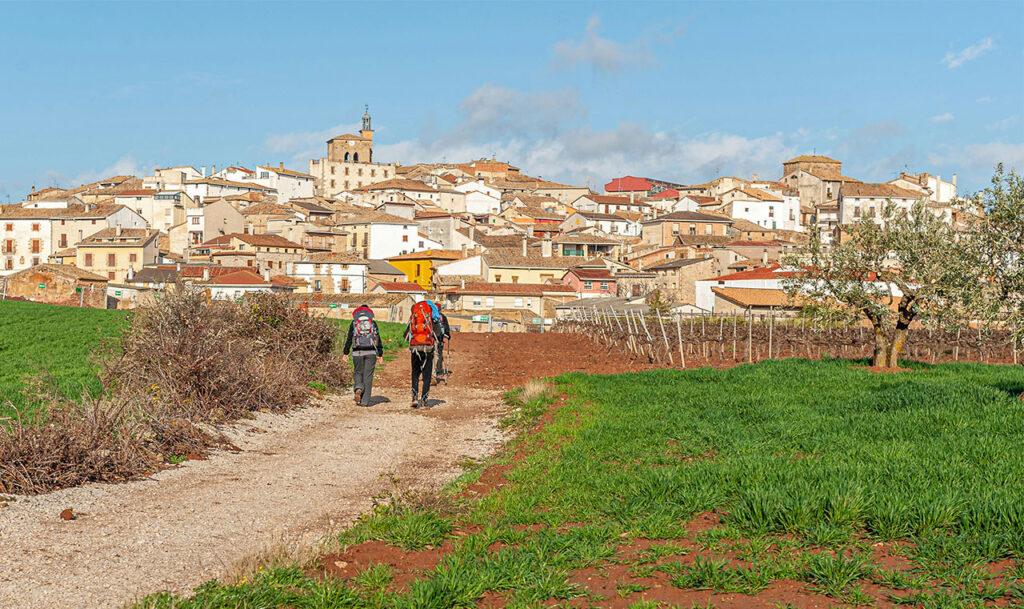 Two pilgrims with large backpacks walking on a dirt trail toward the medieval hilltop town of Cirauqui in northern Spain.