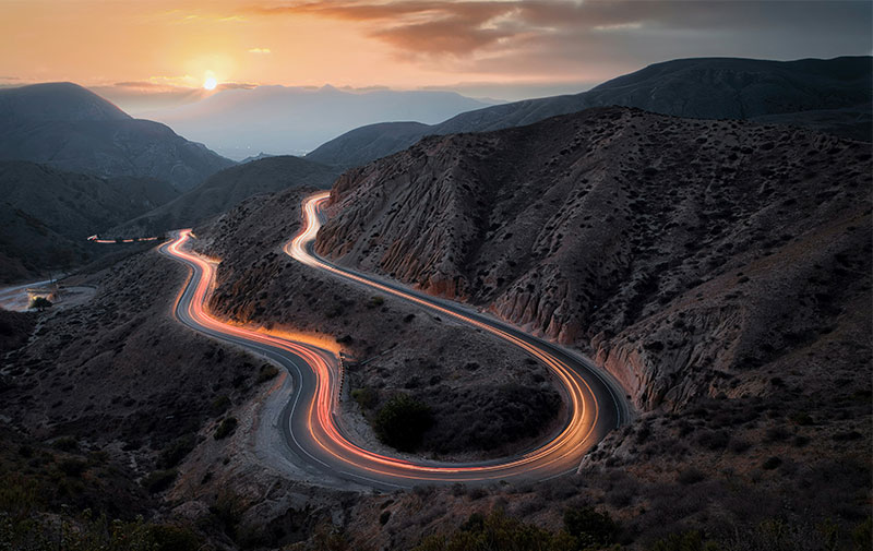 Long exposure showing red and white light trails from cars navigating a steep, winding mountain road in the California desert at sunset.