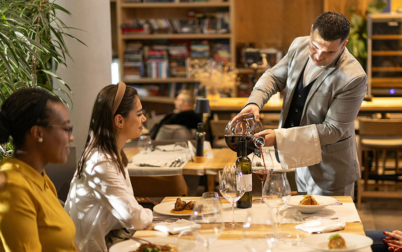 A sommelier in a grey suit pours red wine from a decanter for two women seated at a restaurant table.