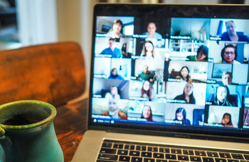 A close-up of a laptop screen during a multi-person video call, set on a wooden table in a Cartagena cafe with a coffee and a smartphone nearby, ready to be used as a personal hotspot.