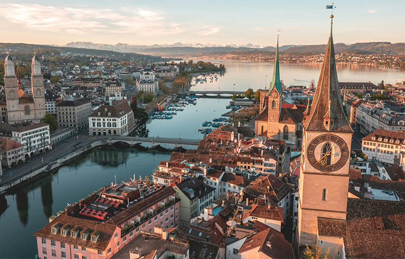 A high-angle, scenic view of Zürich, Switzerland, featuring the Limmat River flowing through the historic center, with the iconic Grossmünster and Fraumünster church spires rising above the traditional tiled rooftops under a soft, hazy sky.