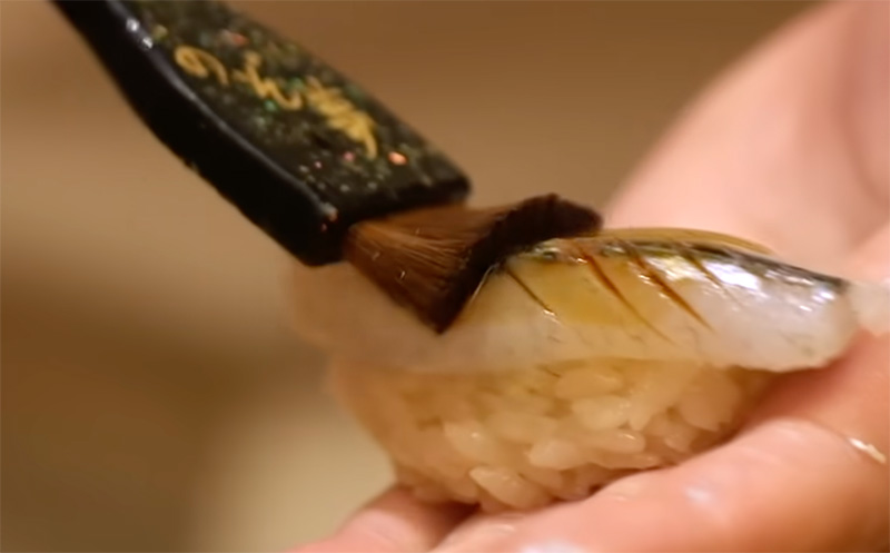 A close-up of a Japanese sushi chef using a traditional brush to lightly apply a thin layer of nikiri soy sauce onto a fresh piece of nigiri sushi.