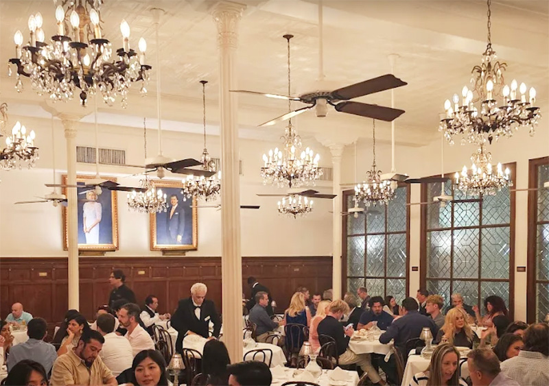 An elegantly dressed crowd dining in the historic Arnaud’s Restaurant in New Orleans, featuring high ceilings, ornate chandeliers, white tablecloths, and large arched windows reflecting the classic French Quarter architecture