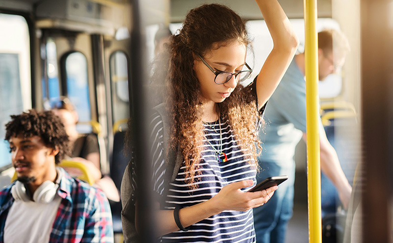 A young woman wearing glasses and a denim jacket stands on a city street, looking down at her smartphone while holding a small black bag.