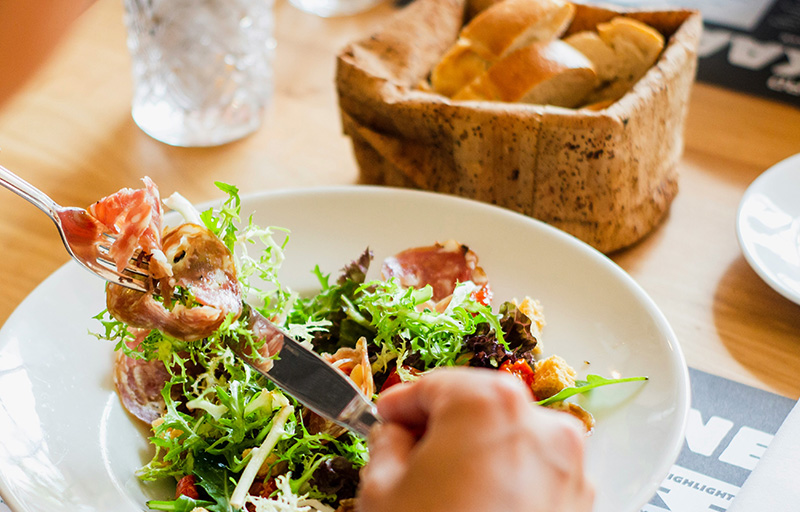 A close-up shot of a plate of food at a restaurant, featuring a toasted sandwich and a fresh green salad, with a person’s hands using a knife and fork in the foregroun