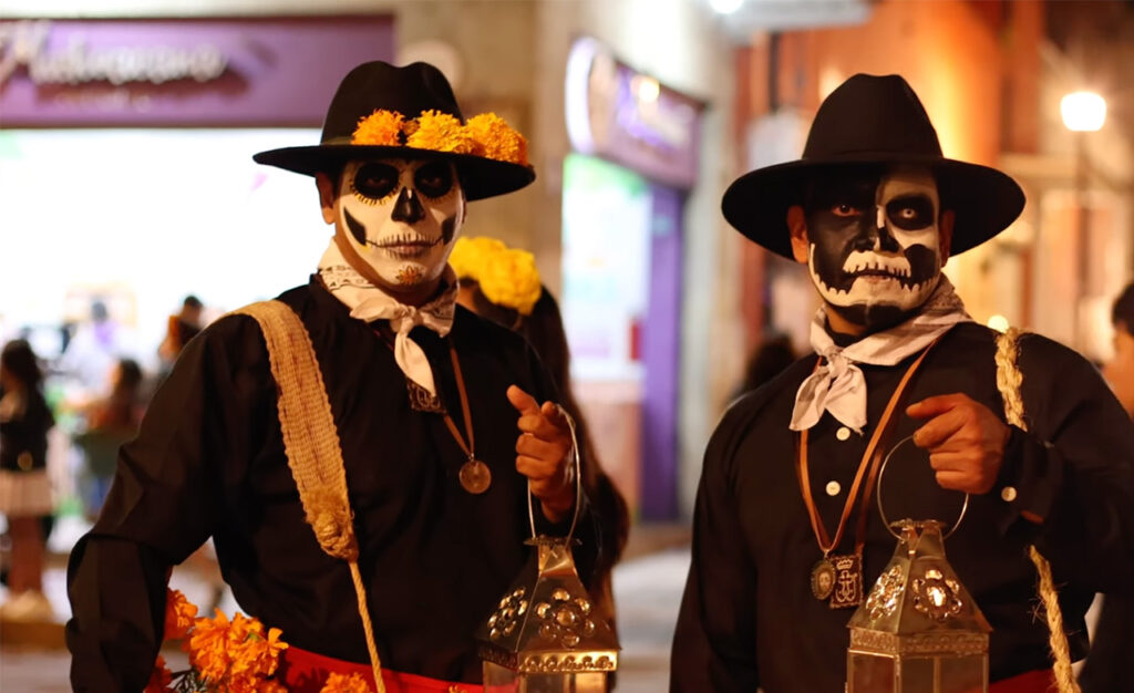 Two people in Oaxaca dressed in elaborate, traditional-style "Día de los Muertos" skeleton face paint and black costumes, posing on a cobblestone street for tourist photos.