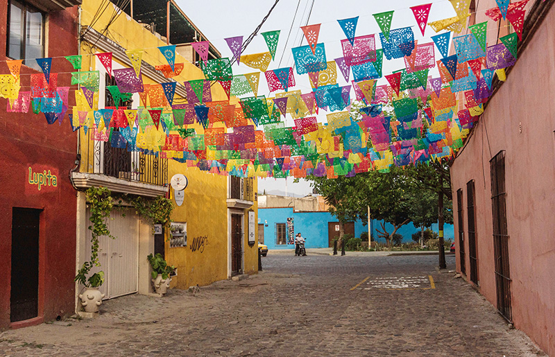 A narrow, cobblestone street in the Jalatlaco neighborhood of Oaxaca, lined with colorful buildings and crisscrossed overhead by rows of vibrant, multi-colored decorative flags (papel picado).