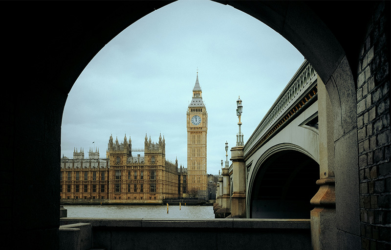 A view of Big Ben and the Elizabeth Tower in London through a stone archway, with the Palace of Westminster and a street lamp in the foreground