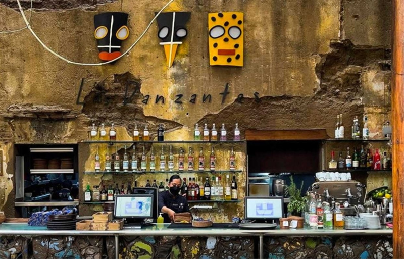 A warm and inviting interior of a traditional mezcalería in Oaxaca's Centro Histórico, featuring a rustic wooden bar lined with various artisanal mezcal bottles and hand-labeled glass jugs (garrafones)