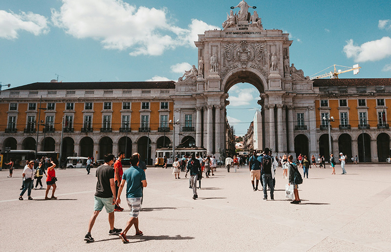 People walking through the historic Praça do Comércio in Lisbon, Portugal, featuring the yellow buildings and the Rua Augusta Arch under a clear blue sky.
