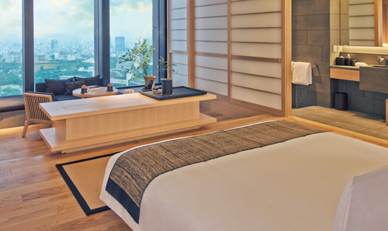 A luxurious, minimalist hotel room at the Aman Tokyo featuring floor-to-ceiling windows that offer a panoramic view of the Tokyo skyline and the Imperial Palace Gardens, with a traditional grey stone soaking tub positioned near the glass.