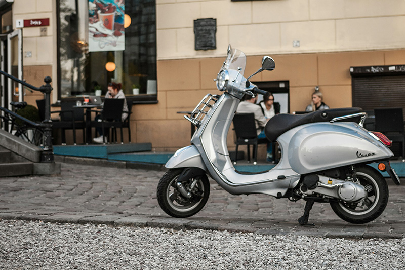 A classic silver Vespa scooter parked on a narrow cobblestone street in an Italian city. Historical buildings with wooden shutters and a small cafe with outdoor seating are visible in the background under soft sunlight.