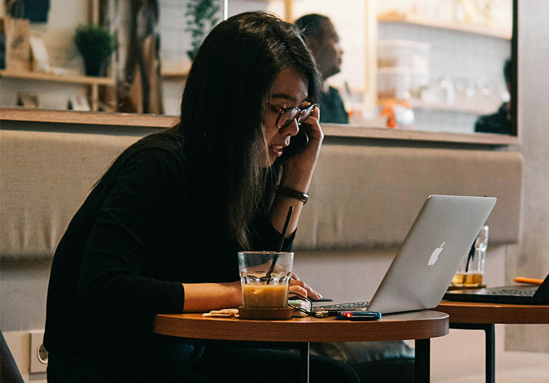 A solo female traveler sitting in a brightly lit cafe, working on a laptop with a focused expression while holding a phone to her ear.