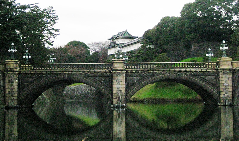 A view of the stone Nijubashi Bridge with its two arches reflecting in the water, leading toward the white buildings and emerald roofs of the Tokyo Imperial Palace surrounded by lush green trees.