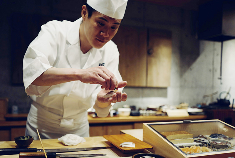 A close-up shot of a sushi chef in a traditional white uniform and cap, meticulously shaping a piece of nigiri sushi by hand at a clean, light-wood counter in a Tokyo restaurant.