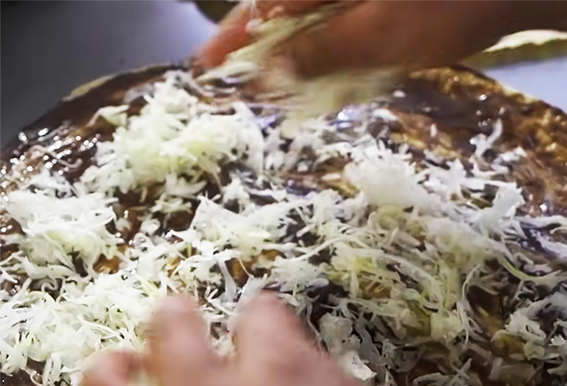 A top-down view of a large, crisp Oaxacan tlayuda spread with black bean paste and topped with a generous layer of shredded quesillo, sliced cabbage, and fresh avocad