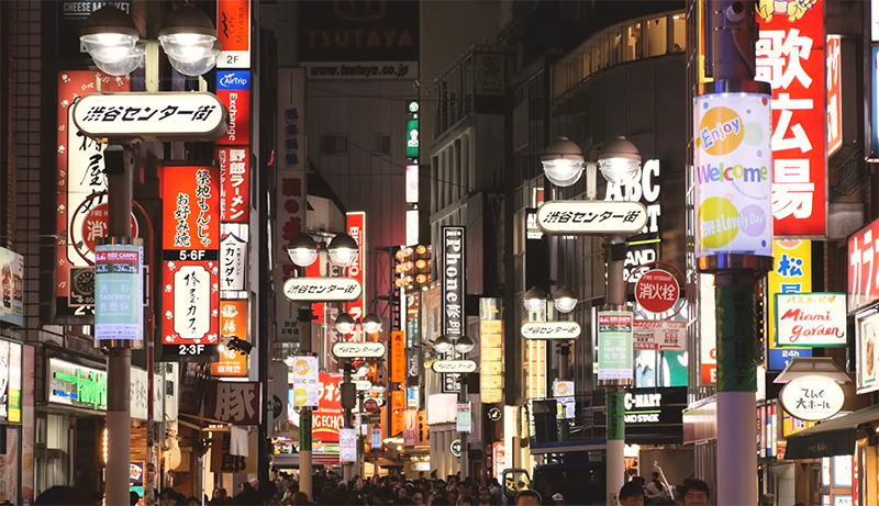 A vibrant, narrow pedestrian street in Shibuya, Tokyo at night, filled with glowing neon restaurant signs, overhead lanterns, and people exploring the local eateries.