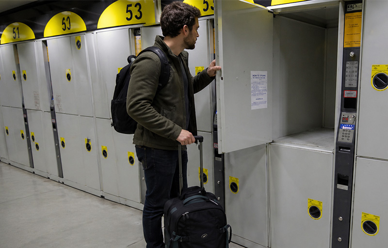 A man in a jacket standing in a bright, modern transit station. He is placing a black backpack into one of many grey and yellow electronic luggage lockers that line the wall.