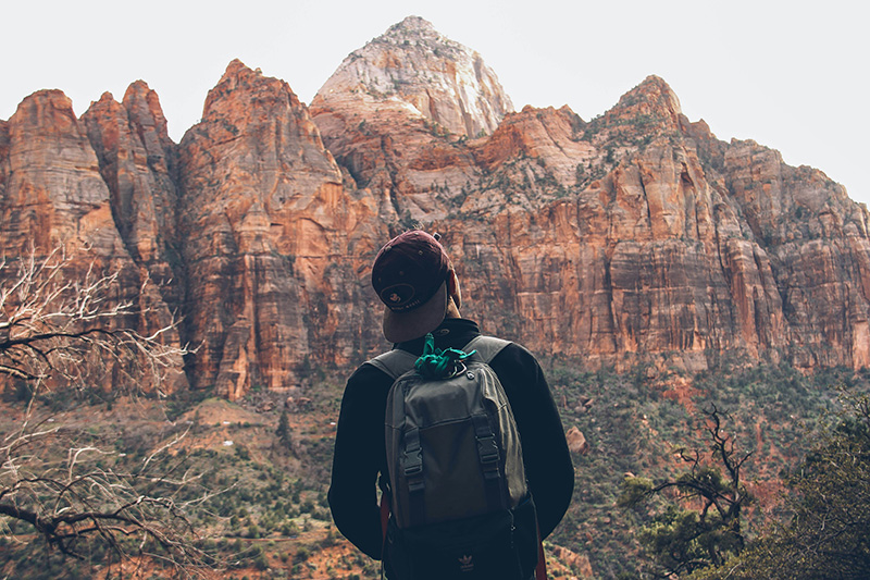 A male traveler seen from behind with a dark backpack, looking out at a rugged mountain range with prominent, jagged rocky peaks and scree slopes.