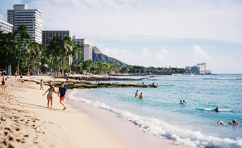 A group of people relaxing on the shore of a tropical beach, with turquoise water and a city skyline visible in the background under a clear sky.