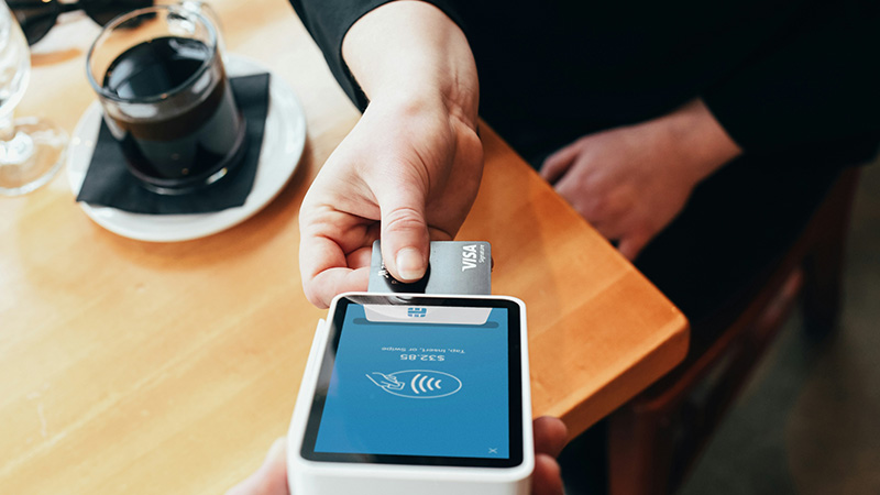 A close-up shot of a hand tapping a credit card on a mobile payment terminal to pay a restaurant bill, with a cup of coffee and a receipt in the background.
