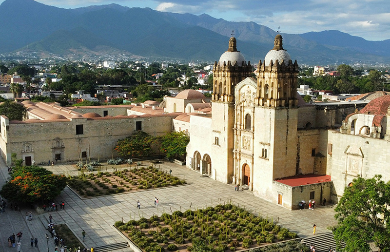 A grand view of the Oaxaca Cathedral (Catedral de Nuestra Señora de la Asunción) in the Centro Histórico, featuring its ornate green quarry stone facade and bell towers under a bright blue sky.